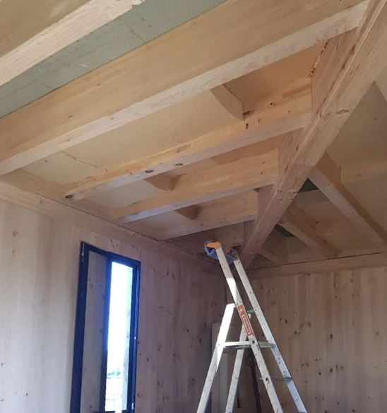 Intérieur de construction en bois avec charpente apparente, murs en panneaux de bois massif, échelle aluminium positionnée au centre