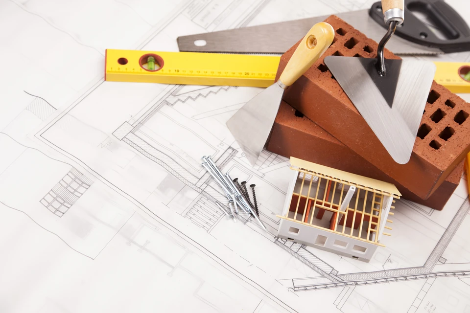 Scène d'intérieur montrant un espace de travail d'architecte ou maçon avec plans techniques étalés, maquette de maison à ossature bois, briques perforées empilées, niveau à bulle jaune et truelle à manche en bois