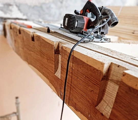 Artisan travaillant en extérieur sur chantier de construction, utilisant un rabot manuel pour façonner une planche en bois